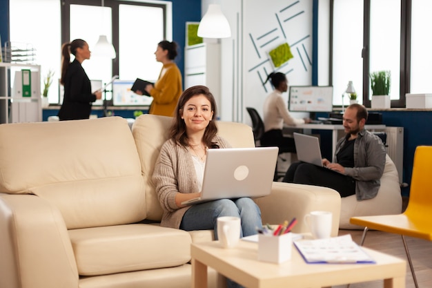 Porträt eines Unternehmers, der auf einem Laptop tippt und lächelnd in die Kamera schaut, während ein vielfältiges Team im Hintergrund arbeitet