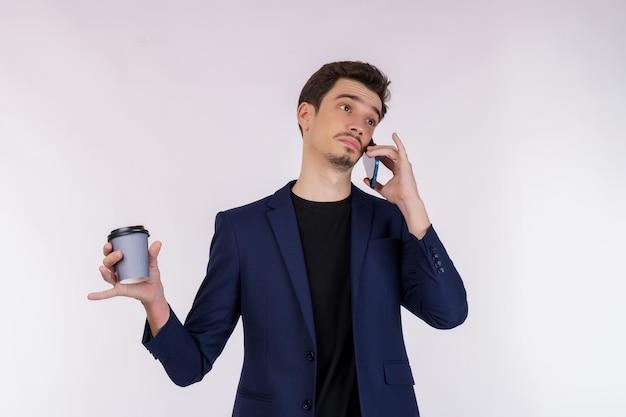 Porträt eines unzufriedenen, gutaussehenden Geschäftsmannes, der per Handy spricht und heißen Kaffee isoliert über weißem Hintergrund hält