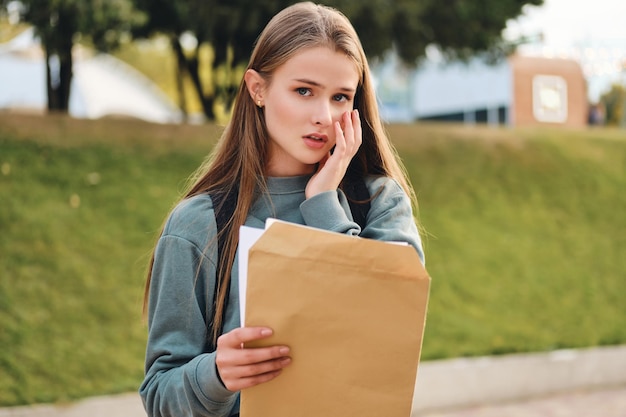 Porträt eines verärgerten, zufälligen Studentenmädchens, das einen Umschlag mit Prüfungsergebnissen öffnet, die traurig in die Kamera im Stadtpark schauen