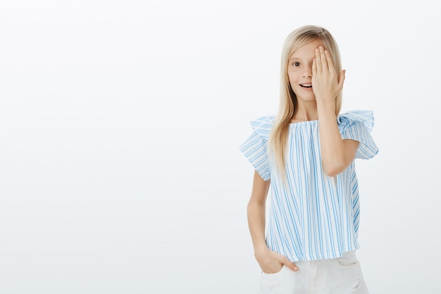 Porträt eines verblüfften freudigen jungen Mädchens mit blonden Haaren in blauer Bluse, das die Hälfte des Gesichts bedeckt und vor Überraschung nach Luft schnappt, mit einem fantastischen Bild betäubt ist und über einer grauen Wand steht