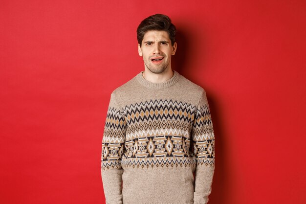 Porträt eines verwirrten erwachsenen Mannes im Weihnachtspullover, der während der Neujahrsferien enttäuscht steht, etwas nicht versteht und auf rotem Hintergrund steht.