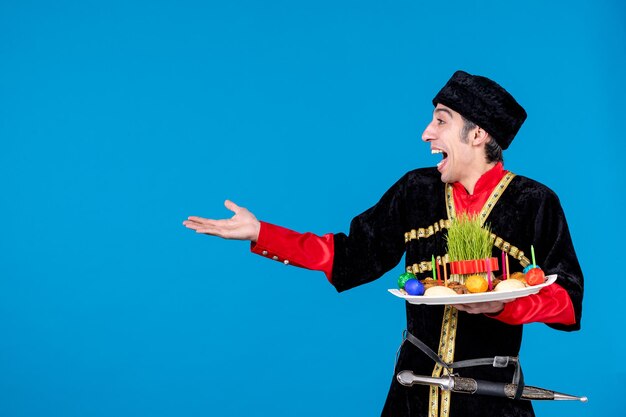 Porträt eines verwirrten jungen Erwachsenen in traditioneller Kleidung, der ein Tablett zeigt, das mit nationalen Süßwaren gefüllt ist und auf blauem Wellenhintergrund zeigt