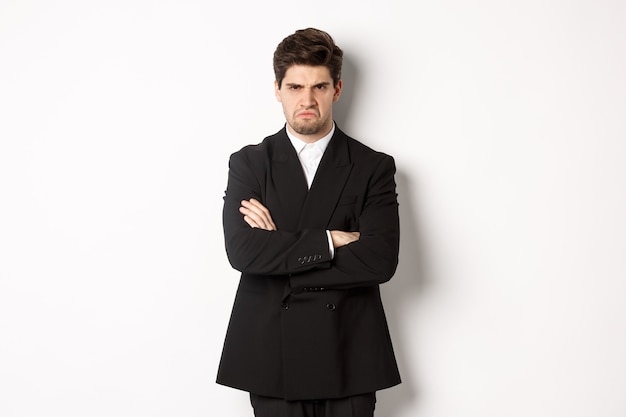 Porträt eines wütenden gutaussehenden Mannes im schwarzen Anzug, die Arme auf der Brust verschränkt und beleidigt aussehend, die Stirn runzelnd und schmollend, wütend auf jemanden, der auf weißem Hintergrund steht