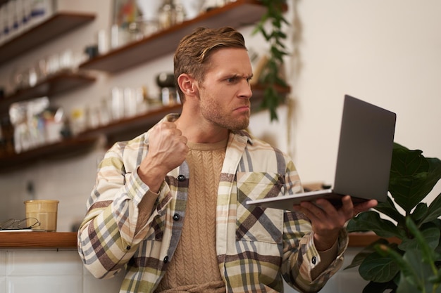 Porträt eines wütenden Mannes in einem Café, der einen Laptop in der Hand hält, auf den Bildschirm starrt und während des Gesprächs eine geballte Faust zeigt