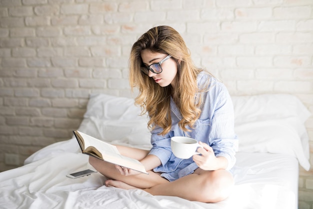 Porträt eines wunderschönen jungen nerdigen Mädchens mit Brille, das ein Buch liest und Kaffee im Bett trinkt