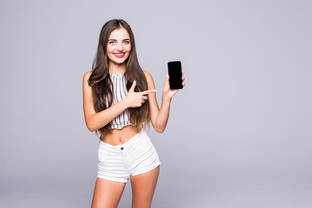 Porträt eines ziemlich freudigen Teenager-Mädchens gekleidet in Jeansjacke, das Finger auf Handy des leeren Bildschirms über grauem Hintergrund zeigt