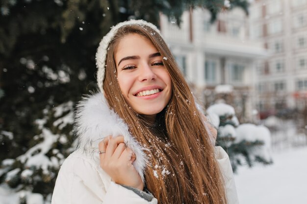 Porträt erstaunliche freudige Frau mit langen brünetten Haaren im Schnee, die Winterzeit auf Straße genießen. Helle Emotionen, gute Laune, Lächeln, Glück, Winterferien.