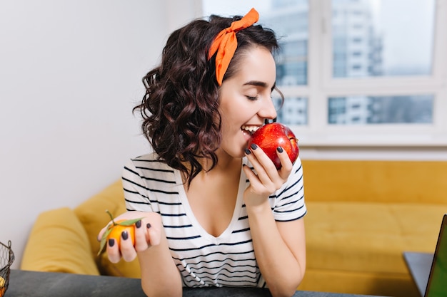 Kostenloses Foto porträt freudige aufgeregte junge frau mit dem lockigen geschnittenen haar, das roten apfel in der modernen wohnung genießt. lächeln, spaß haben, zu hause chillen, gemütlichkeit, entspannung, glück