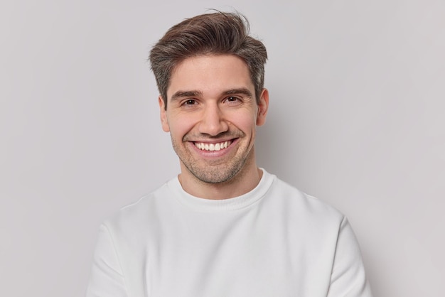 Porträt gutaussehender Mann mit dunklen Haarborsten und zahnigem Lächeln in weißem Sweatshirt fühlt sich sehr froh, Posen im Innenbereich zu haben Zufriedener Europäer, der gute Laune hat, lächelt positiv Emotionskonzept