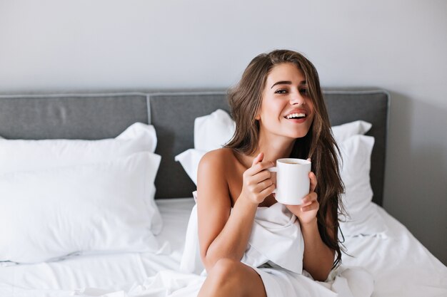 Porträt hübsches Mädchen mit frechen Schultern, die Kaffee auf Bett am Morgen in der Wohnung trinken.