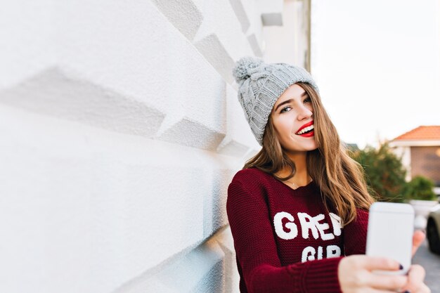 Porträt hübsches Mädchen mit langen Haaren im Marsala-Pullover, der Selfie auf grauer Wand macht. Sie trägt eine Strickmütze und hat rote Lippen.