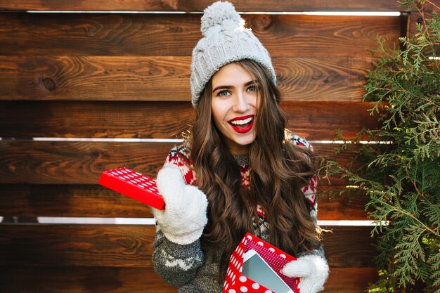 Porträt hübsches Mädchen mit langen Haaren und roten Lippen mit Weihnachtsbox auf Holz. Sie trägt eine Strickmütze, Handschuhe und lächelt.