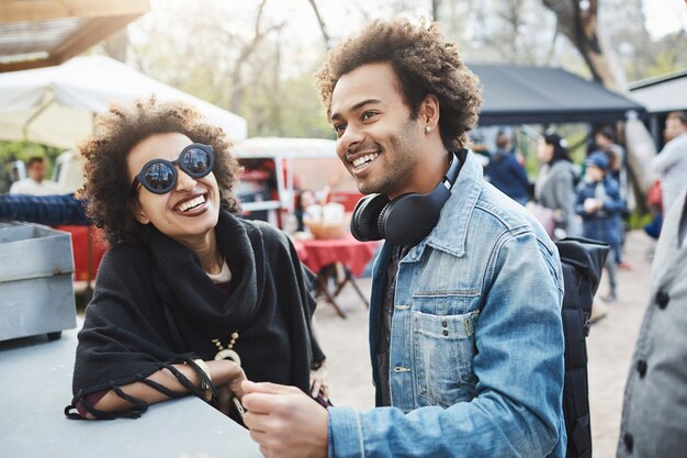 Porträt im Freien eines glücklichen afroamerikanischen Paares mit Afro-Frisuren, das sich auf Tisch während des Lebensmittelfestivals stützt, Zeit zusammen verbringt und auf ihre Bestellung wartet.