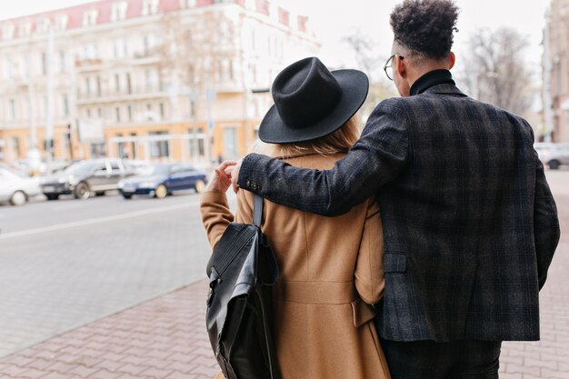 Porträt im Freien von der Rückseite des afrikanischen Mannes im karierten Anzug, der sanft blonde Freundin umarmt. Mädchen im beige Mantel, der Stadtansichten während des Datums genießt.