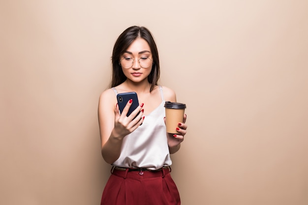 Porträt in voller Länge der lächelnden asiatischen Frau unter Verwendung des Mobiltelefons beim Halten der Tasse Kaffee, um über beige Wand isoliert zu gehen