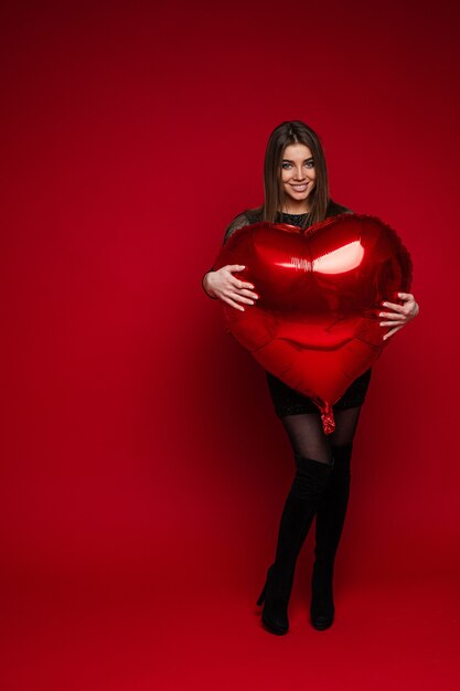 Porträt in voller Länge des fröhlichen brünetten Mädchens im Kleid und in den Stiefeln, die roten herzförmigen Ballon auf rotem Hintergrund umarmen. Saint Valentine Konzept.