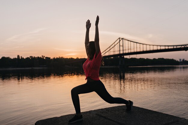 Porträt in voller Länge des glücklichen schlanken Mädchens, das Yoga auf Natur tut. Außenaufnahme der erfreuten Dame, die das Training am Abend genießt.