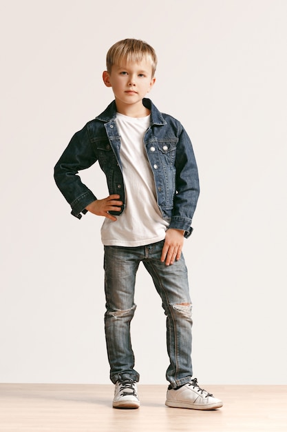 Porträt in voller Länge des niedlichen kleinen Jungen in stilvollen Jeans-Kleidern und lächelnd, auf weiß stehend. Kindermode-Konzept