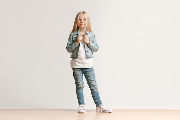 Porträt in voller Länge des niedlichen kleinen Kindermädchens in der stilvollen Jeanskleidung, die Kamera betrachtet und lächelt und gegen weiße Studiowand steht. Kindermode-Konzept