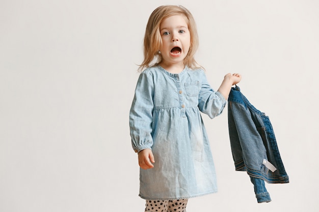 Porträt in voller Länge des niedlichen kleinen Kindermädchens in der stilvollen Jeanskleidung und lächelnd, auf weiß stehend. Kindermode-Konzept