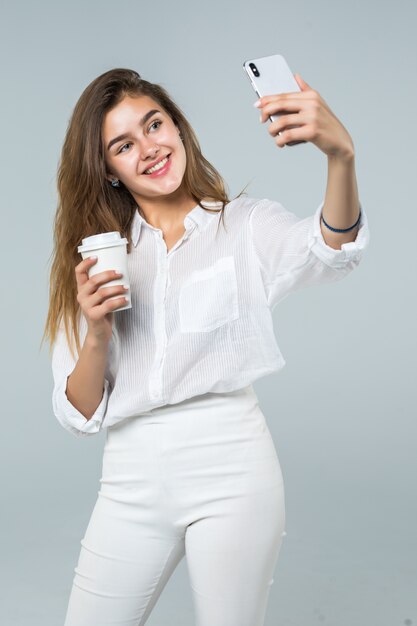 Porträt in voller Länge eines glücklichen lächelnden Mädchens unter Verwendung des Mobiltelefons beim Stehen und Halten der Kaffeetasse über weißem Hintergrund