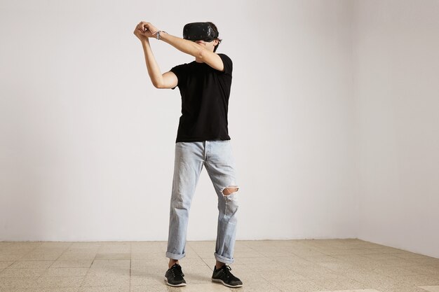 Porträt in voller Länge eines jungen kaukasischen Modells in hellblauen zerrissenen Jeans und schwarzem T-Shirt, das Baseball oder Tennis in VR-Brille spielt