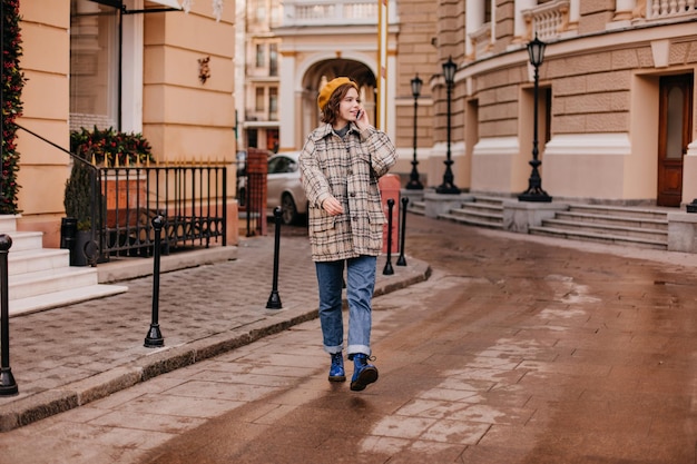 Porträt in voller Länge von Dame in stylischen Jeans und übergroßem Mantel vor dem Hintergrund der schönen europäischen Straße Mädchen mit kurzen lockigen Haaren spricht per Telefon