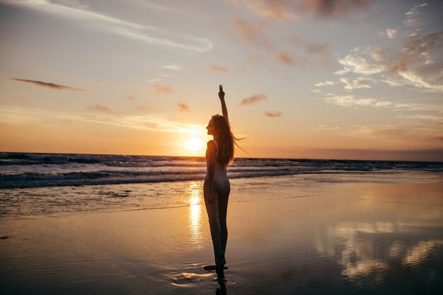 Porträt in voller Länge von der Rückseite des Mädchens, das den Sonnenuntergang im Meer betrachtet. Außenaufnahme des erfreuten weiblichen Modells, das am Abend an der Ozeanküste kühlt.