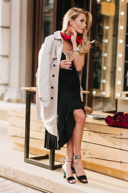 Porträt in voller Länge von hübscher blonder Frau, die neben Restaurant mit Glas Wein sitzt und gutes Wetter genießt. Foto im Freien des Mädchens im schwarzen Kleid, das Champagner allein trinkt.