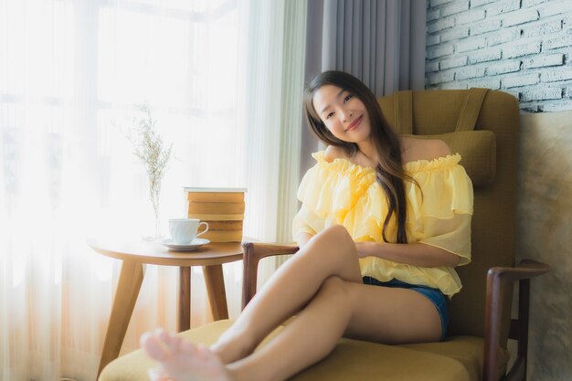 Porträt junge asiatische Frau sitzen auf Sofastuhl und lesen Buch mit Kaffeetasse