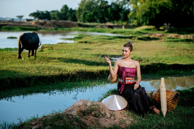 Porträt junge hübsche asiatische frau in schöner thailändischer traditioneller kleidung am reisfeld, sie sitzt in der nähe von angelausrüstung