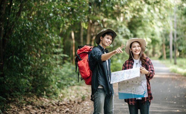 Porträt junger asiatischer gutaussehender mann mit rucksack und trekking-hut und hübscher freundin, die auf der papierkarte steht und die richtung überprüft, während sie auf dem waldweg geht, rucksackreisekonzept