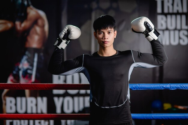 Porträt junger gutaussehender Mann in Sportkleidung und weißen Boxhandschuhen, die die Arme heben, um im Fitnessstudio auf der Leinwand zu posieren, gesunder Mann Training Boxklasse