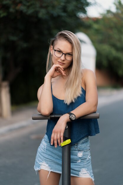 Porträt junges schönes Mädchen in der Brille, die einen Elektroroller im Sommer auf der Straße reitet