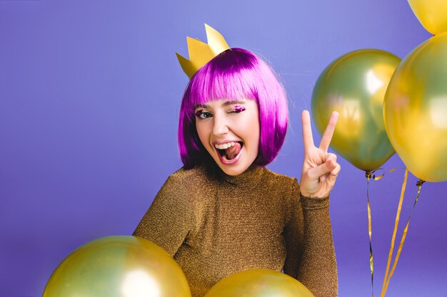 Porträt lustige junge Frau mit lila Haarschnitt Spaß haben. Goldene Luftballons umgeben, zeigen Zunge, drücken positive Gesichtsgefühle aus, Krone auf Kopf, Geburtstagsfeier.