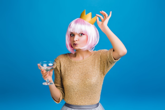 Porträt lustige lustige stilvolle junge Frau im goldenen Pullover, rosa Haarschnitt, Krone auf Kopf. Spaß haben, Champagner trinken, Neujahrsparty feiern, Geburtstag.