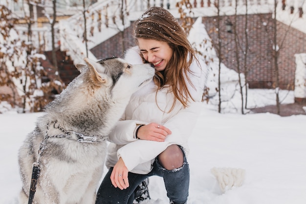 Porträt niedliche schöne Momente des Husky-Hundes, der modische junge Frau im Freien im Schnee küsst. Fröhliche Stimmung, Winterferien, Schneezeit, echte Freundschaft, Tierliebe.