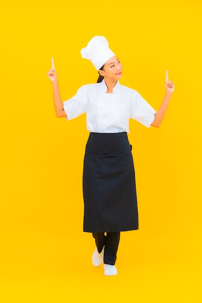 Porträt schöne junge asiatische Frau in Koch- oder Kochuniform mit Hut auf gelbem isoliertem Hintergrund