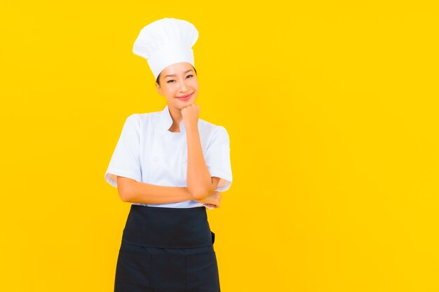 Porträt schöne junge asiatische Frau in Koch- oder Kochuniform mit Hut auf gelbem isoliertem Hintergrund