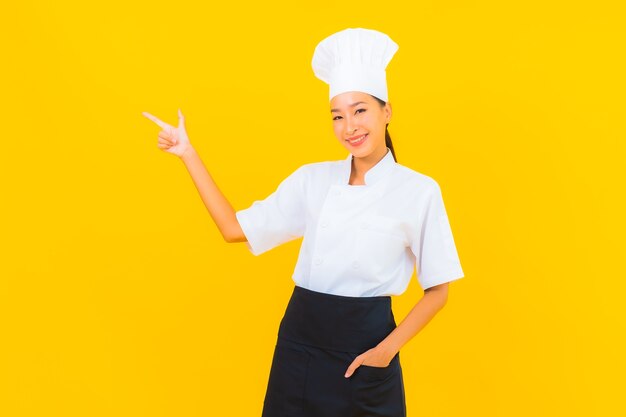 Porträt schöne junge asiatische Frau in Koch- oder Kochuniform mit Hut auf gelbem isoliertem Hintergrund