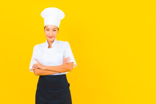 Porträt schöne junge asiatische Frau in Koch- oder Kochuniform mit Hut auf gelbem isoliertem Hintergrund