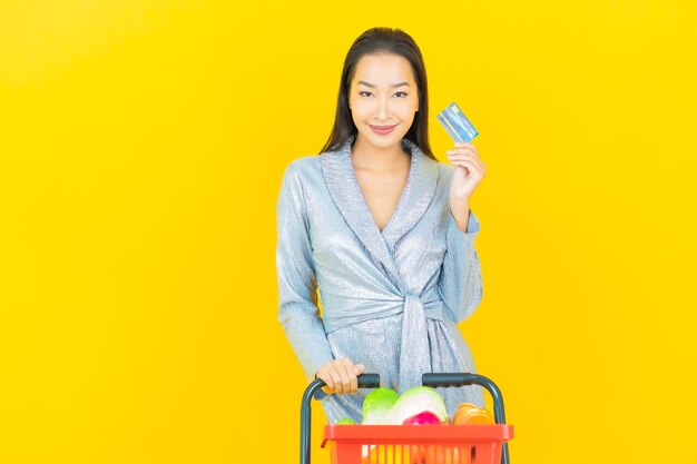 Porträt schöne junge asiatische Frau lächeln mit Lebensmittelkorb vom Supermarkt auf gelber Wand
