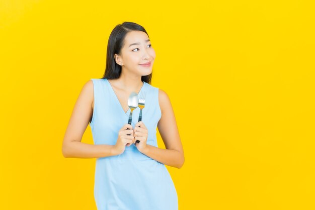 Porträt schöne junge asiatische Frau lächeln mit Löffel und Gabel auf gelber Farbwand
