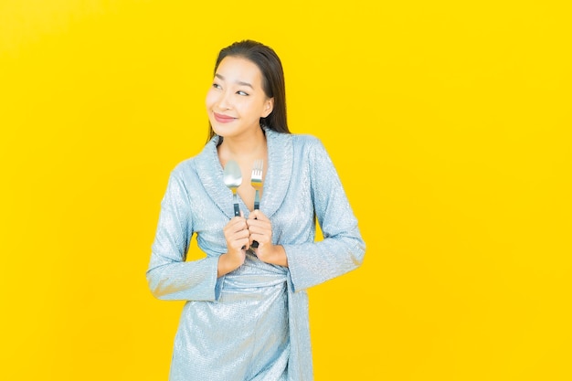 Porträt schöne junge asiatische Frau lächeln mit Löffel und Gabel auf gelber Wand