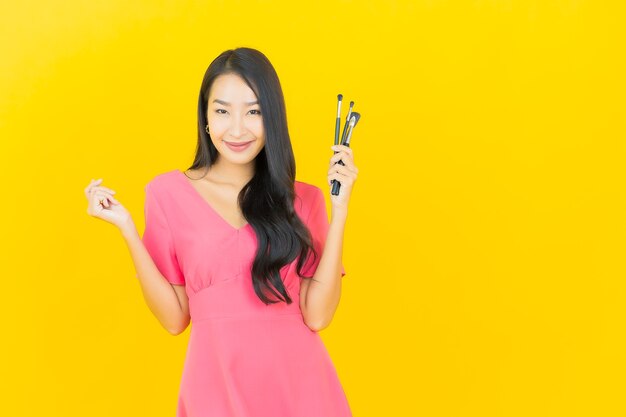 Porträt schöne junge asiatische Frau lächelt mit kosmetischem Make-up Pinsel auf gelber Wand