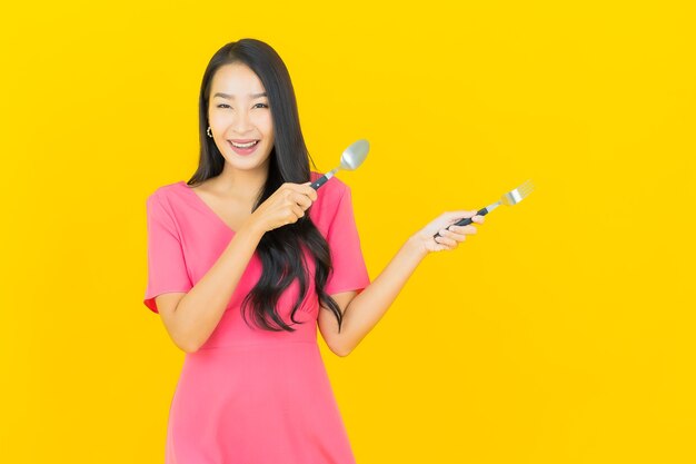 Porträt schöne junge asiatische Frau lächelt mit Löffel und Gabel auf gelber Wand