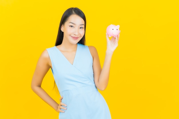 Porträt schöne junge asiatische Frau mit Sparschwein auf gelber Farbwand