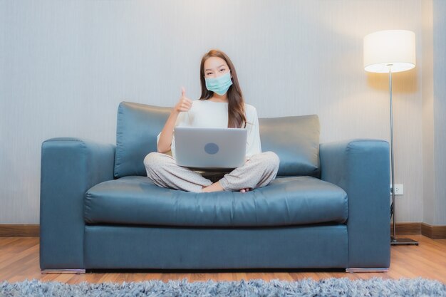 Porträt schöne junge asiatische Frau tragen Maske und verwenden Computer-Laptop auf Sofa im Wohnzimmer Interieur