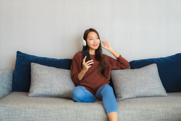 Porträt schöne junge asiatische Frau verwenden Smart-Handy mit Kopfhörer für Musik auf Sofa im Wohnzimmer Interieur zu hören