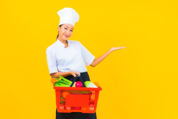 Porträt schöne junge asiatische Kochfrau mit Einkaufskorb aus dem Supermarkt auf gelbem isoliertem Hintergrund